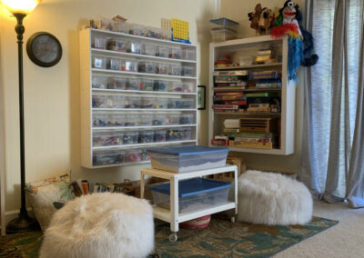 Picture of play area in room 5 at TheraThrive Lafayette, showing two sand trays and shelf of sand tray toys, game shelf, fluffy bean bag chairs, books, puppets, and other toys.
