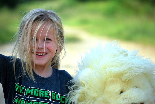 An image of a child smiling looking happy and holding a large stuffed animal, with a background of grass and sandy paths.
